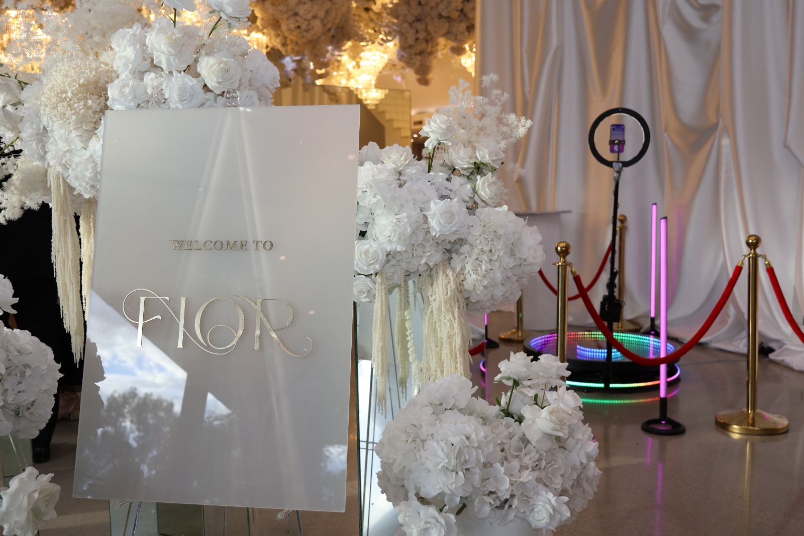 Welcome sign framed by white florals beside a 360 booth