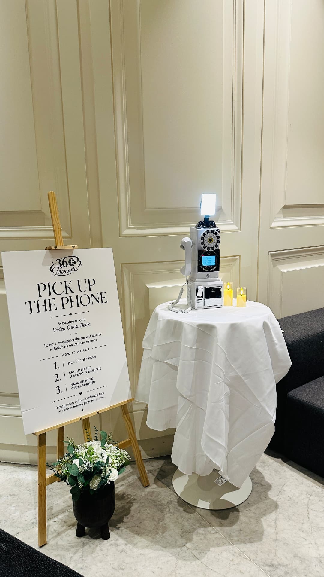 Video guest book on a white draped table beside a Pick Up the Phone sign and flowers
